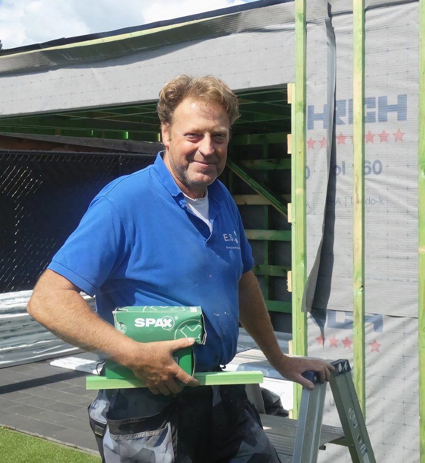 Eric Snoekx in blauwe werkpolo, met een doos schroeven in de hand, voor een houtskelet in opbouw.