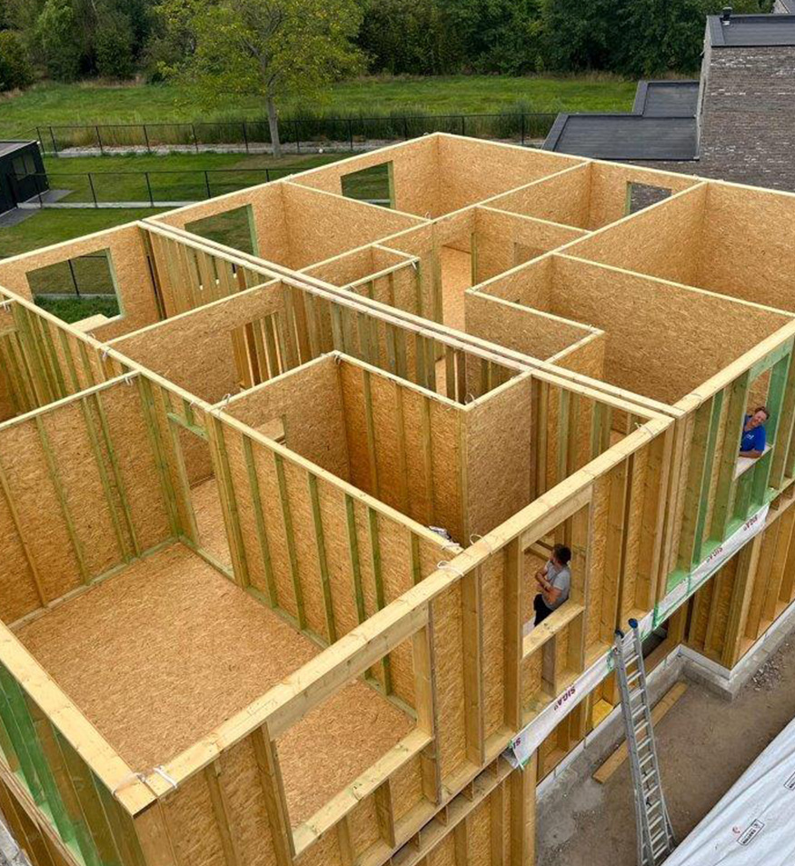 Luchtbeeld van een houtskelet in opbouw: de wanden en kamerindeling van een woning staan overeind, twee vakmensen werken aan de gevel.