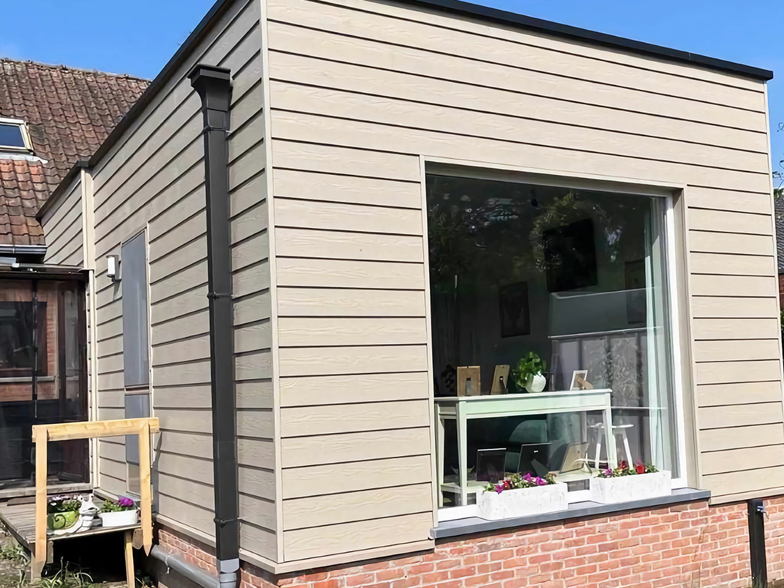 Uitbouw met lichte houten gevelbeplanking en een groot raam, geplaatst tegen een bestaande woning in baksteen.