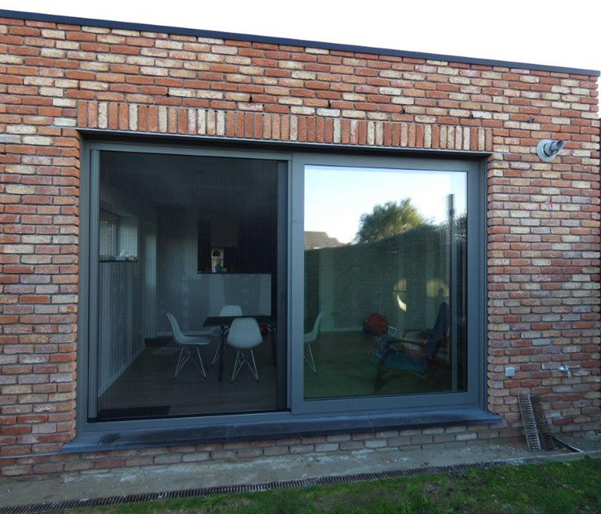 Woonuitbreiding in oude baksteen met een groot schuifraam dat uitkijkt op de tuin.